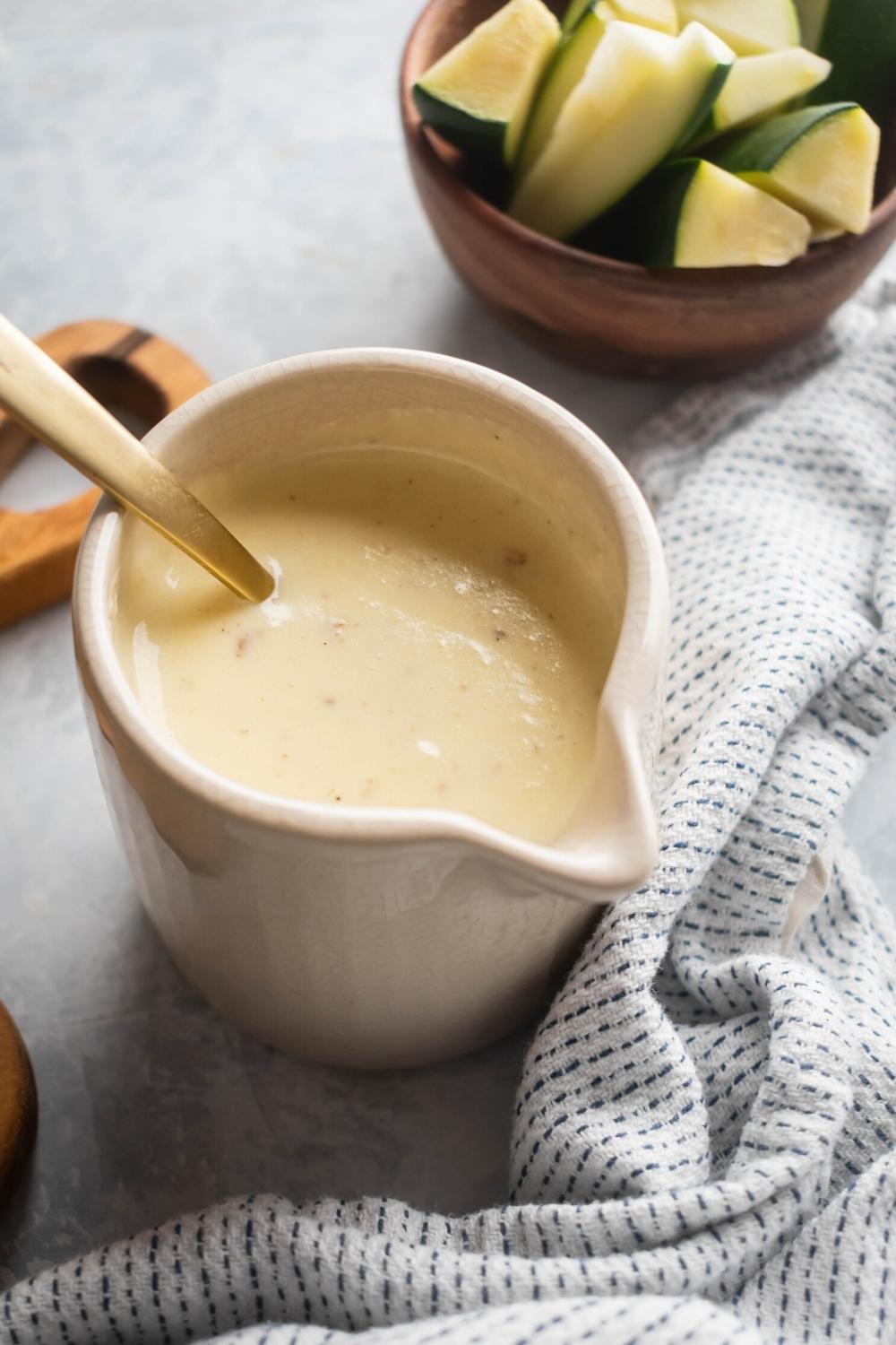 Cheese sauce in a white picture with a gold spoon submerged in it. Behind it is part of a wooden bowl with sliced zucchini in it.