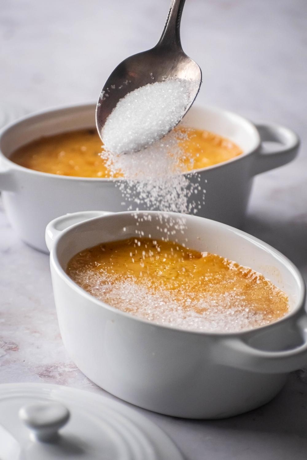 A spoon sprinkling granulated erythritol on top of crème brûlée in a ramekin. There is another ramekin filled with crème brûlée behind it.