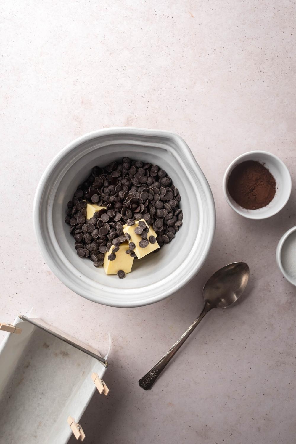 A white bowl with chocolate chips and butter in it. There is a small white bowl filled with cocoa powder next to it.