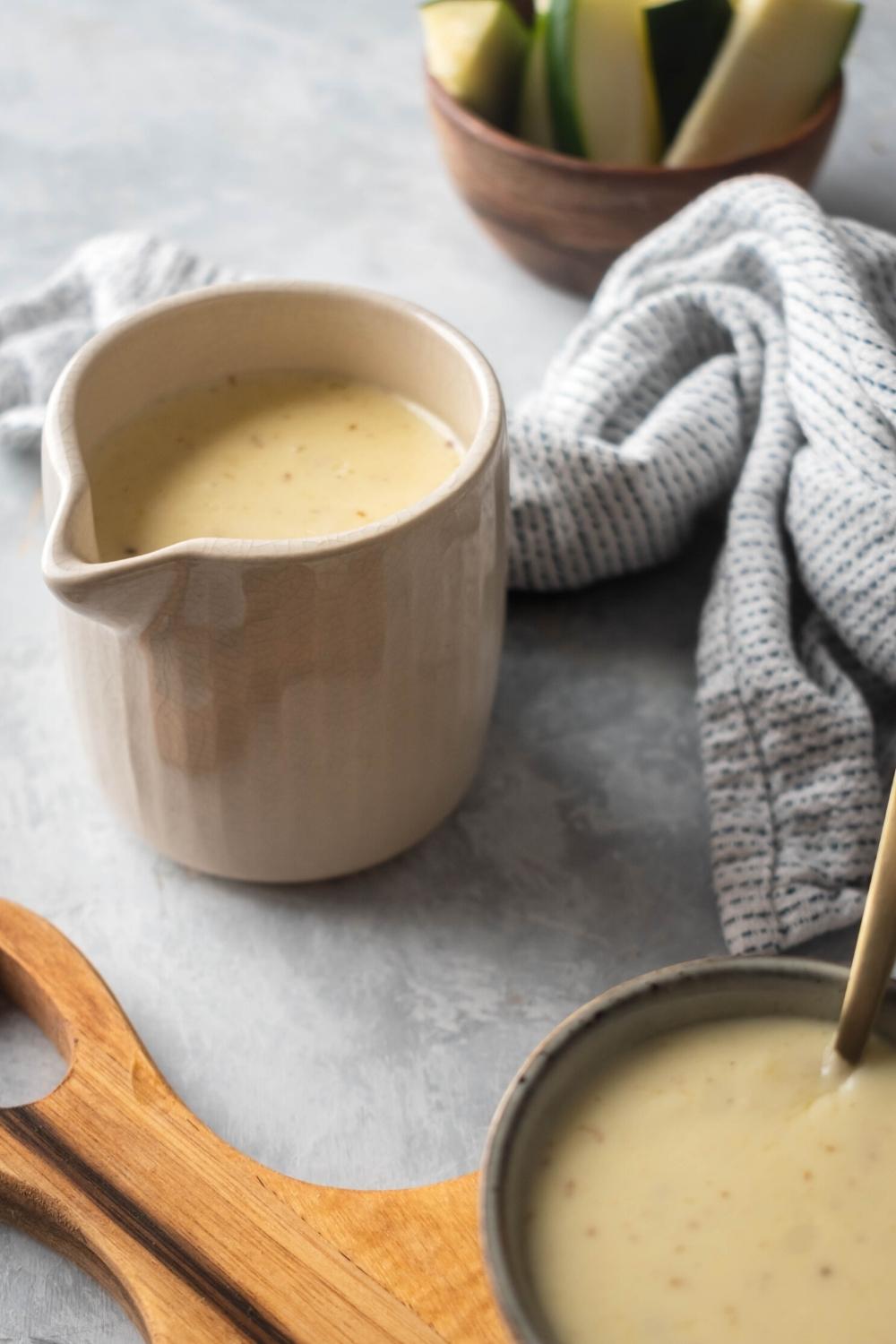 A white pitcher with cheese sauce in it. In front of him part of a wooden cutting board as part of another bowl filled with the cheese sauce.