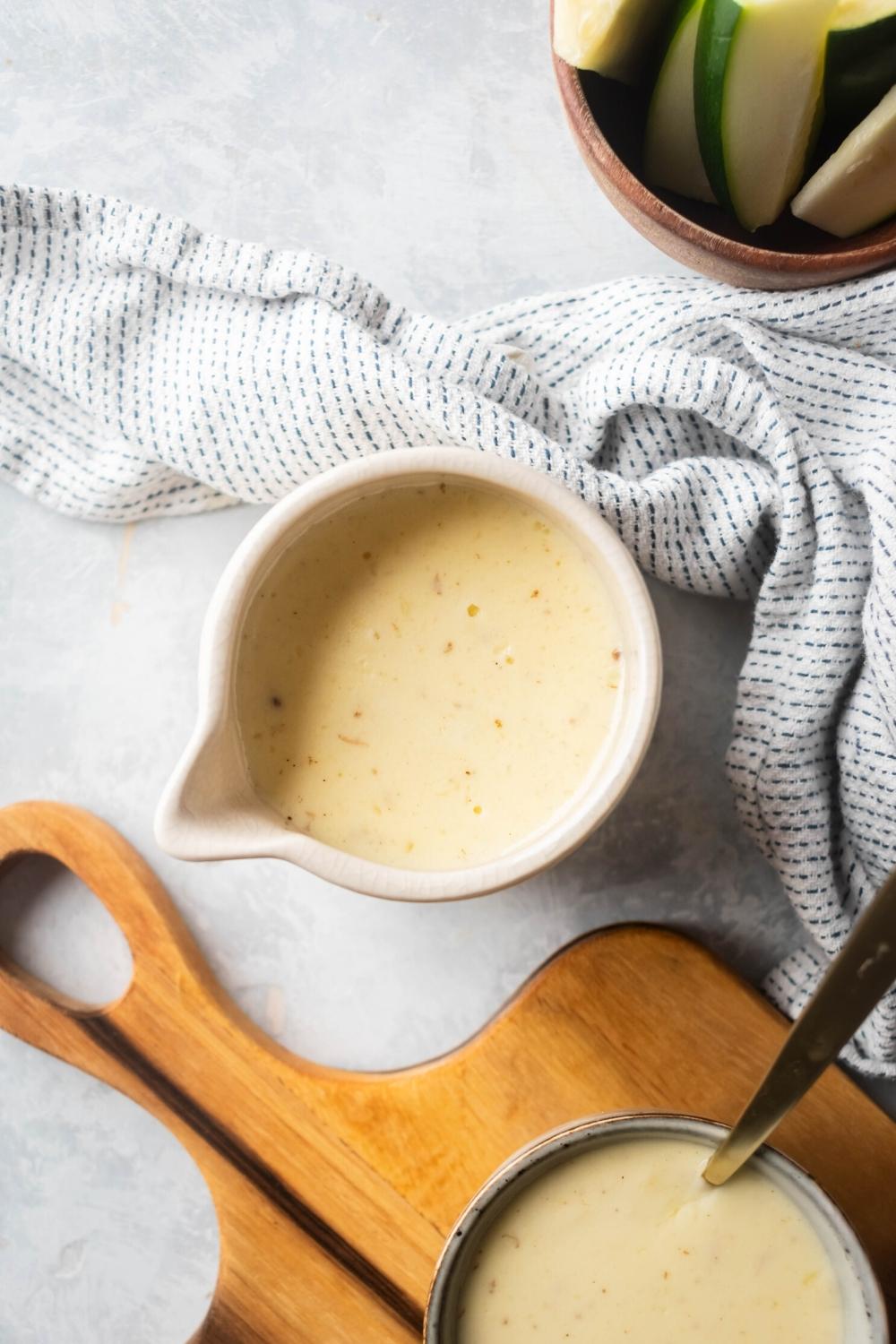 Cheese sauce and white pitcher on top of a gray counter. In front of it is part of a wooden cutting board with part of a bowl of the queso on it and behind it is a towel in behind that part of a bowl with zucchini.