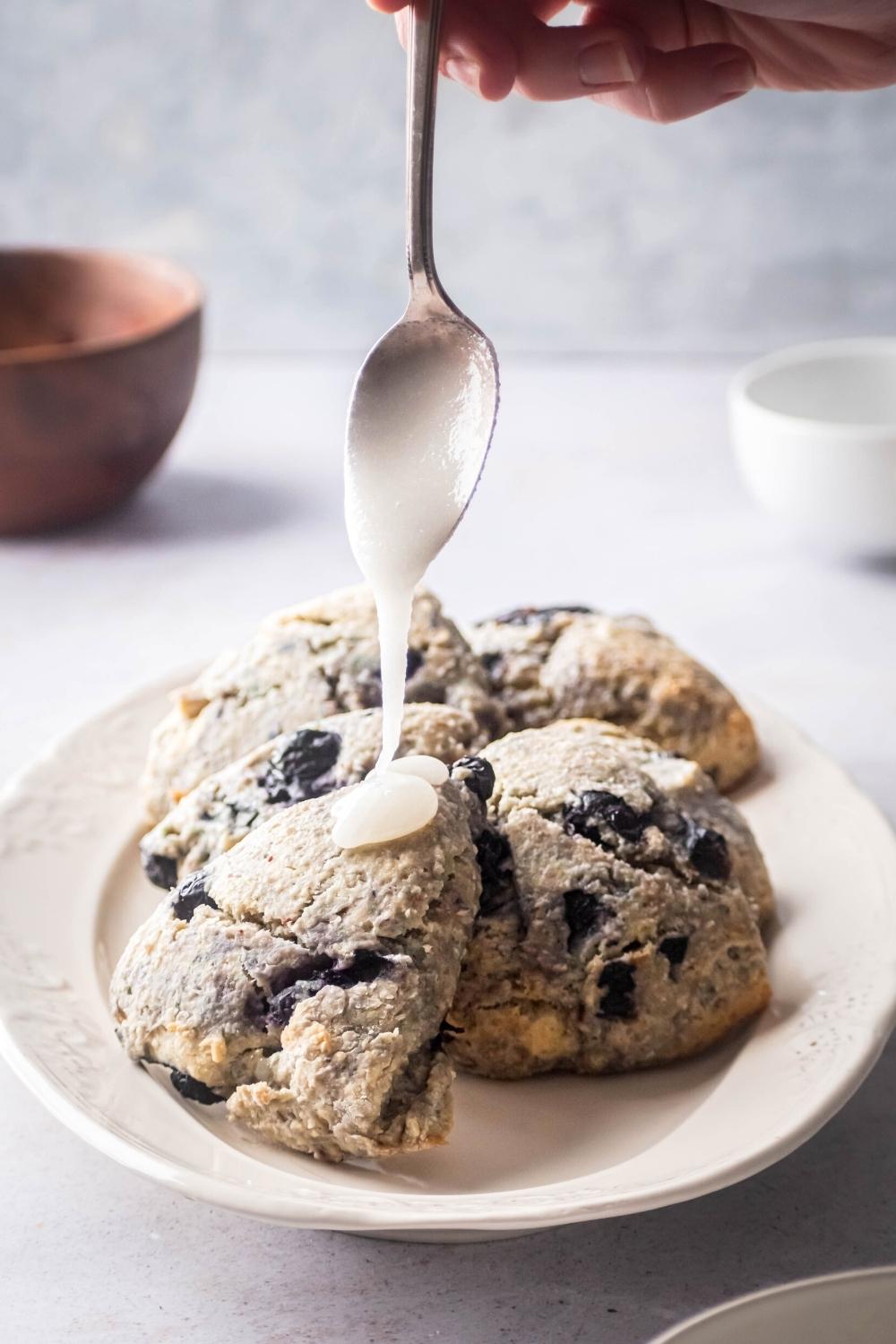 A hand holding a spoon drizzling glaze on top of a blueberry scone. The scone is on a white plate along with a couple of other scones.