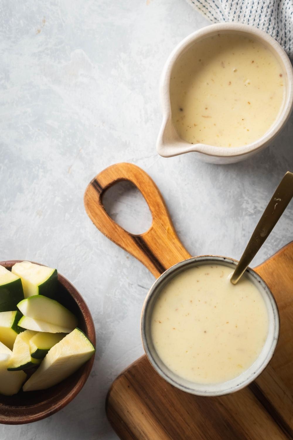 A bowl of cheese sauce with a spoon in it on a wooden cutting board and a gray counter. To the left of it as part of a bowl of slice zucchini into the right is a picture of the cheese sauce.