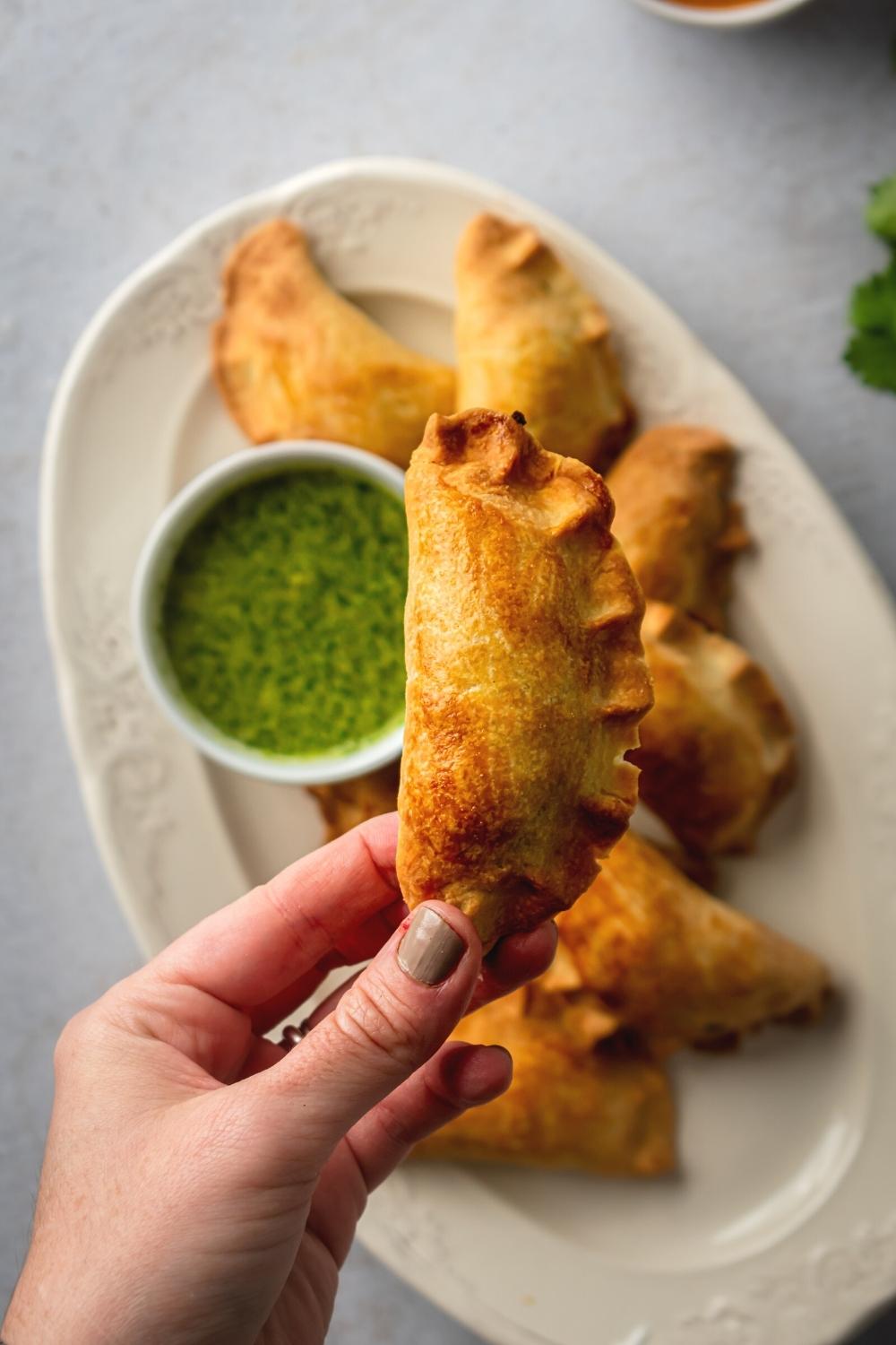 A hand holding a beef empanada over an oval shaped white plate filled with beef empanadas and a small white cup of salsa Verde.