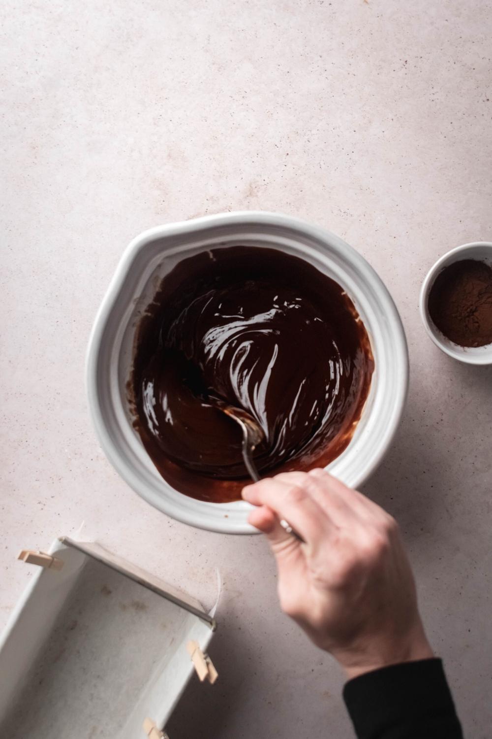 A white bowl filled with melted chocolate with a hand holding a spoon mixing the chocolate.