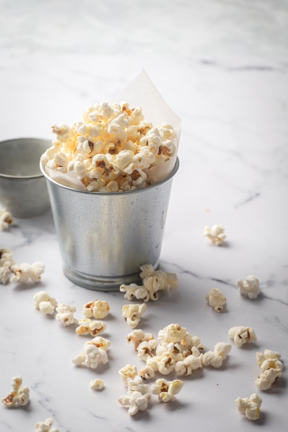 A tin cup filled with popcorn on a grey counter. There is a bunch of popcorn in front on the cup on the counter.