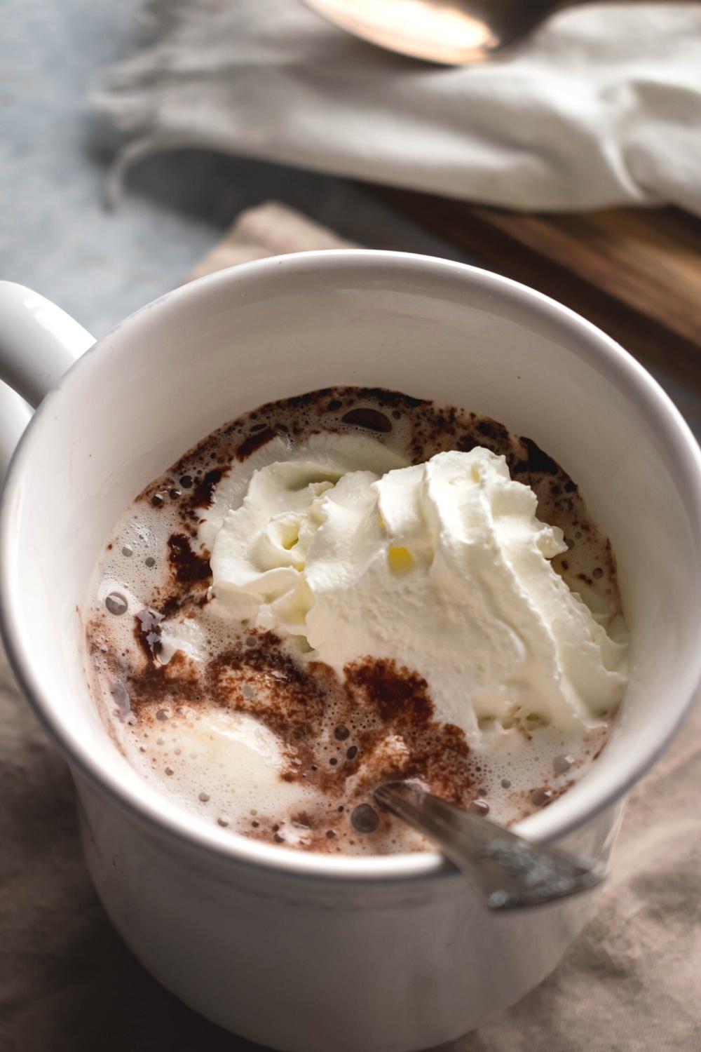 A white mug filled with hot chocolate and some whip cream on top with a spoon submerged in it.