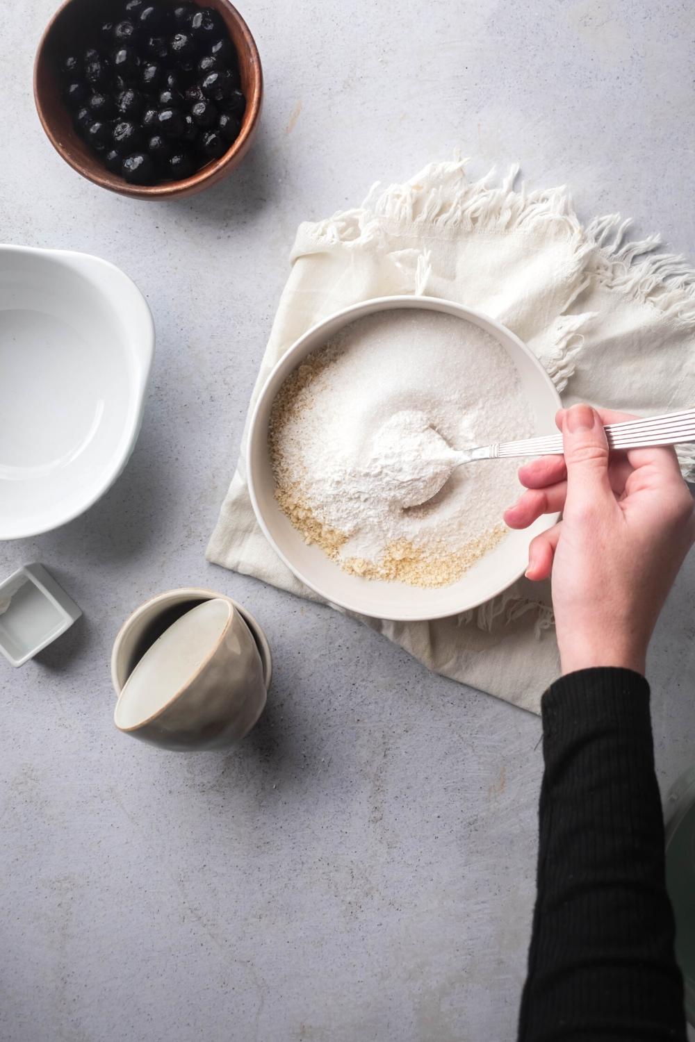 A bowl filled with dry ingredients for scones with a spoon submerged in it being held by a hand. In front of the bowl is a bowl of blueberries.