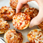 A hand holding a pizza bagel. There are a bunch of other pizza bagels around them.