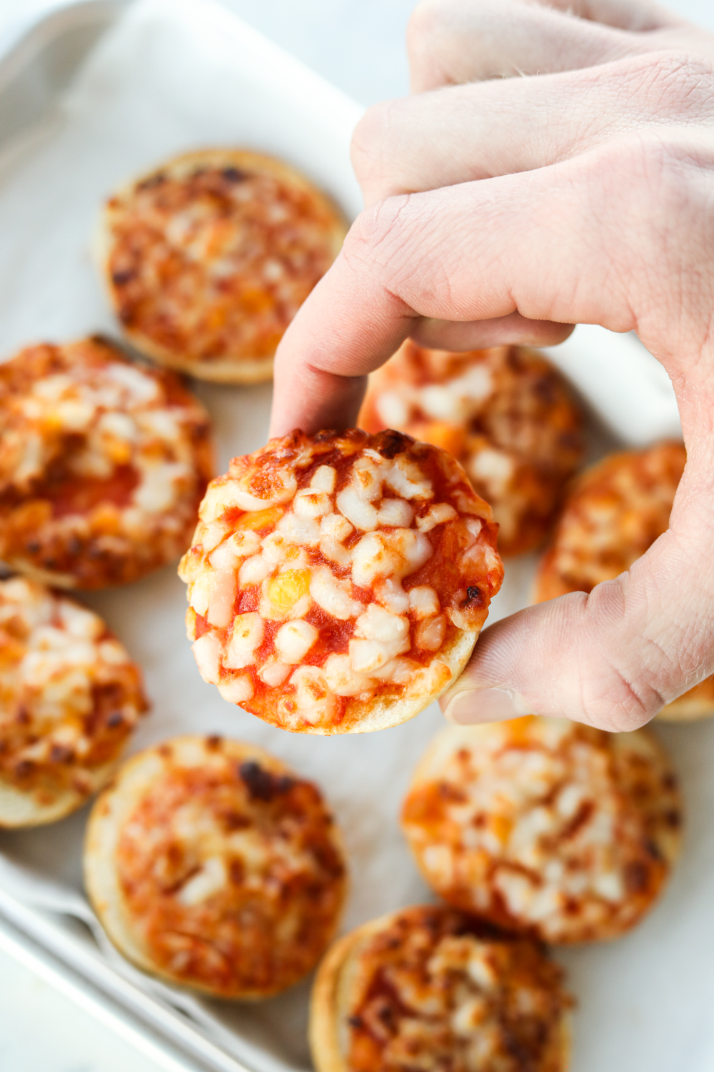 A hand holding a pizza bagel above a bunch of other pizza bagels on a white napkin.