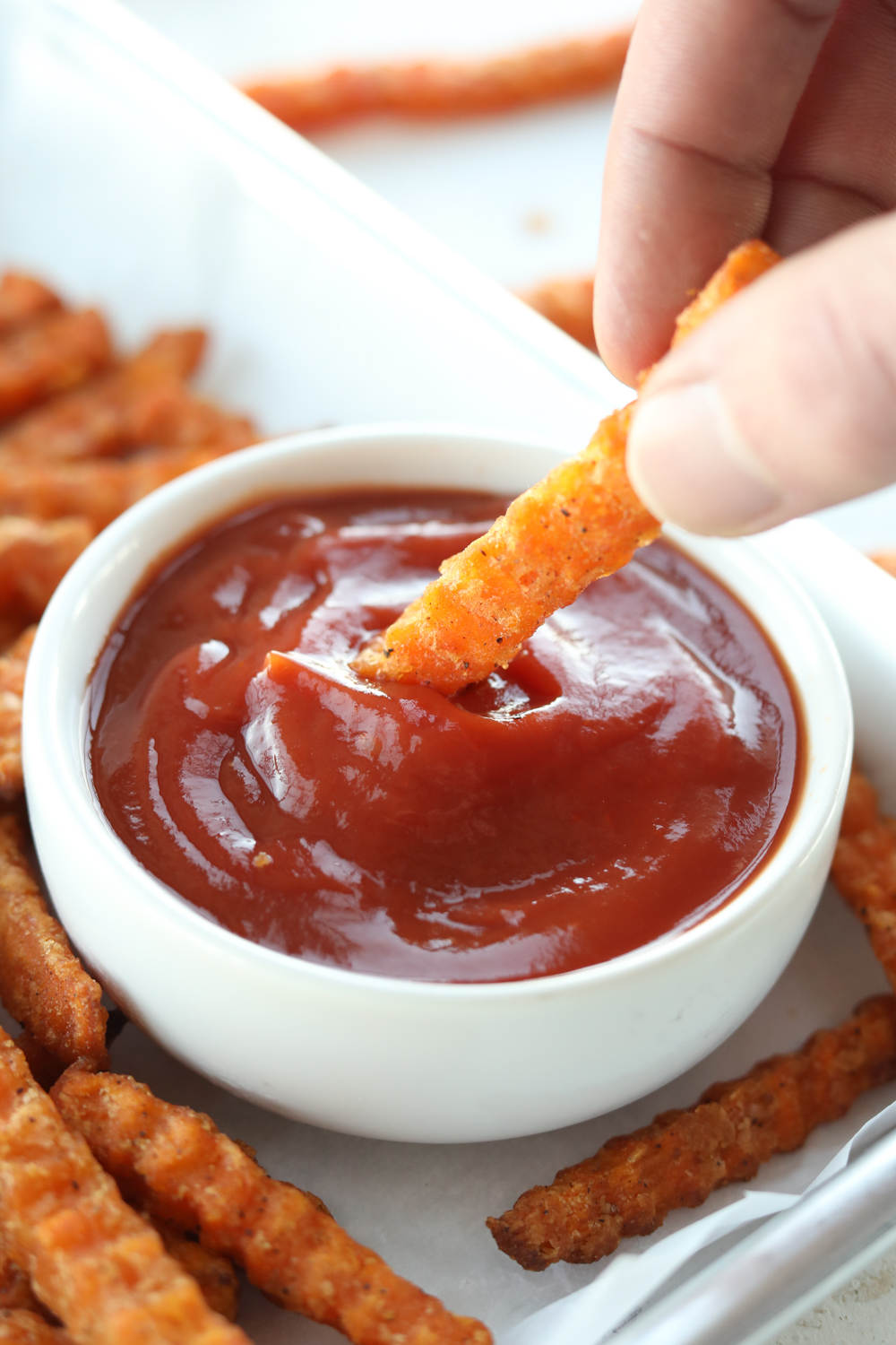 A hand holding a sweet potato fry dipping it into a small cup of catch-up. The catch-up is on a piece of parchment paper and there are a bunch of sweet potato fries around it.