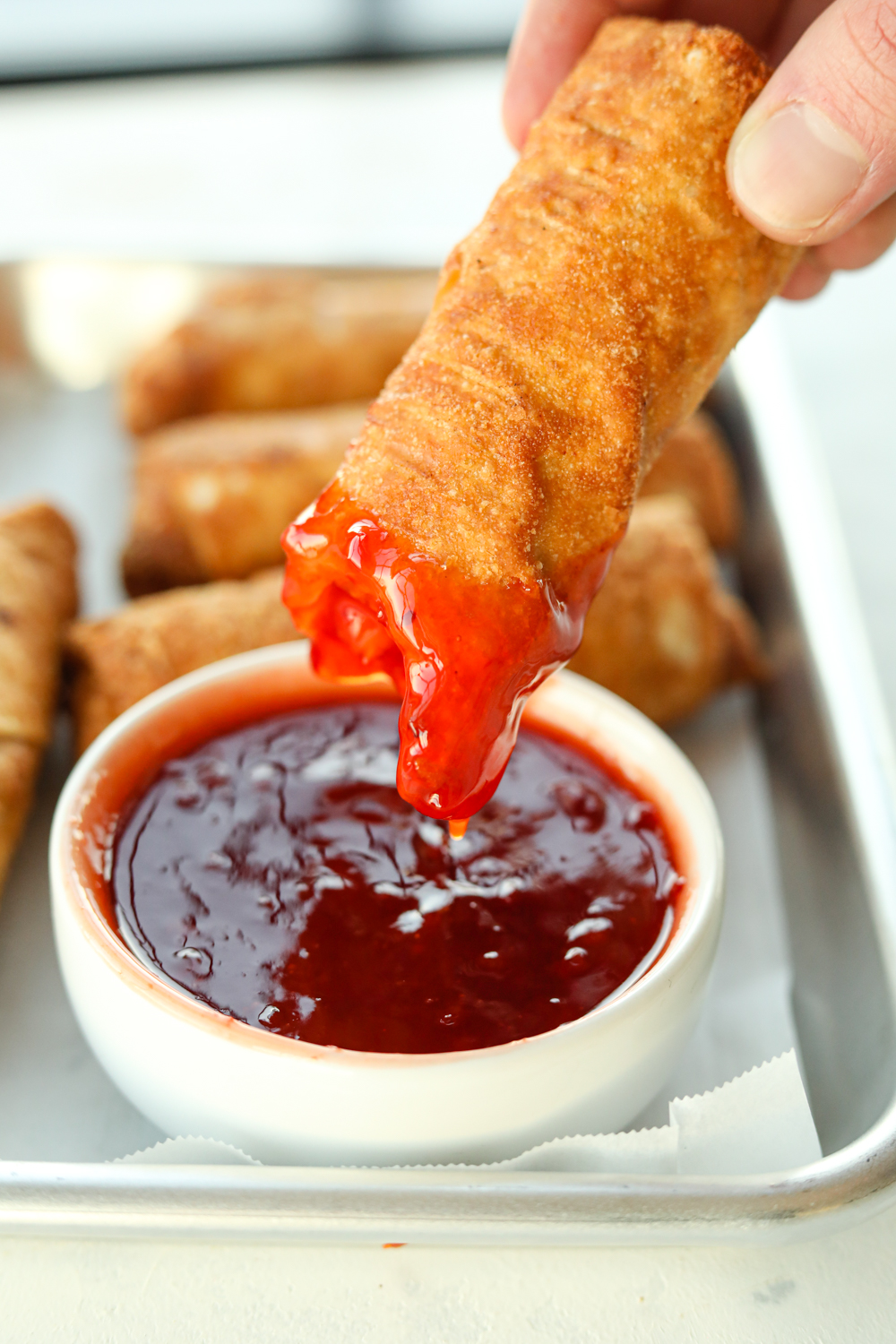 Fingertips holding an eggroll with a bite out of the front of it covered and sweet-and-sour sauce. The eggroll is being held over sweet-and-sour sauce and a white top and behind the cup are three other eggrolls on parchment paper on top and baking sheet.