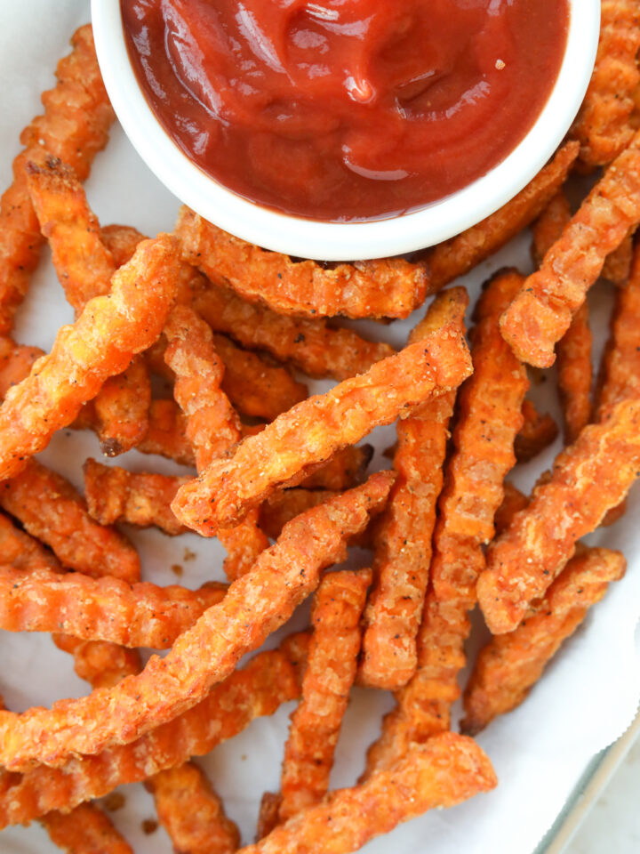 Sweet potato fries on top of one another on a piece of parchment paper. There is part of a cup of ketchup in front of the sweet potato fries on the parchment paper.