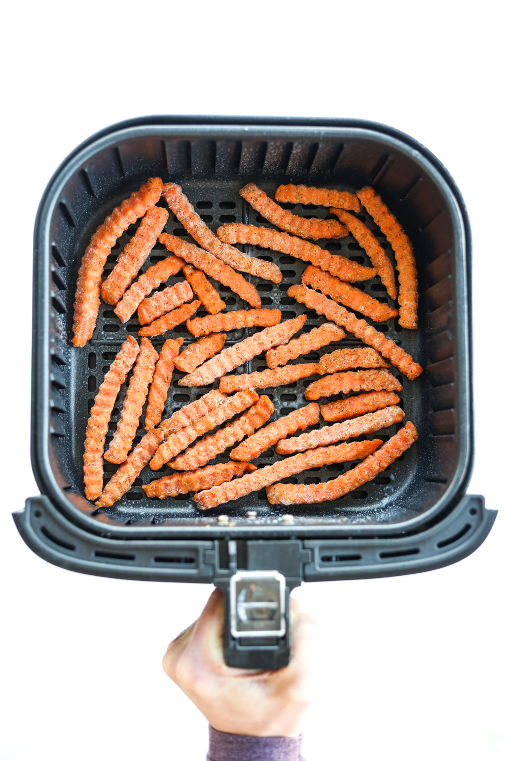 A hand holding an air fryer with a bunch of frozen sweet potato fries in the air fryer basket.
