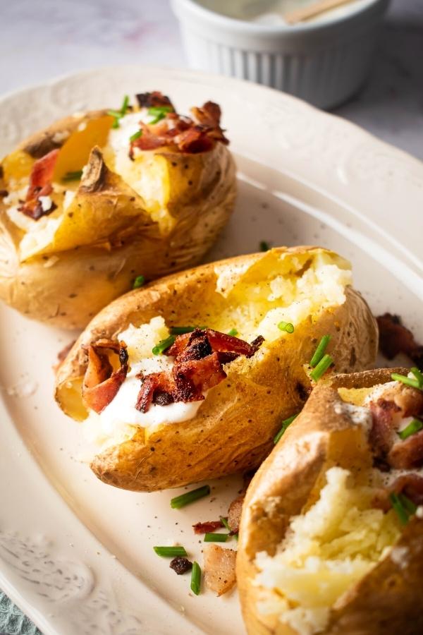 A white plate that has three baked potatoes on it. The baked potatoes are split open and they have some sour cream, chives, and bacon in them.