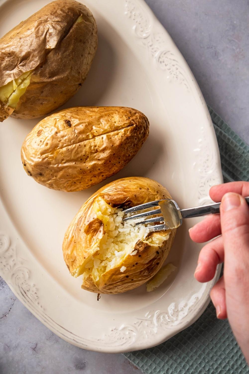 A white plate with three baked potatoes on it. A hand is holding a fork and splitting open the front baked potato.