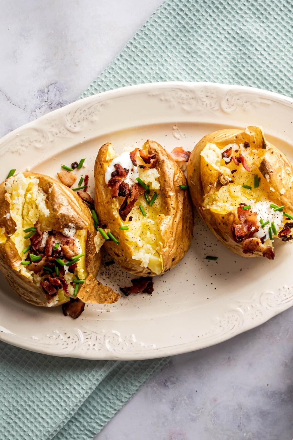 Three baked potatoes next to one another on top of a white plate that is on a blue tablecloth on a white counter. The big potatoes are all split open and filled with chives, bacon, and sour cream.
