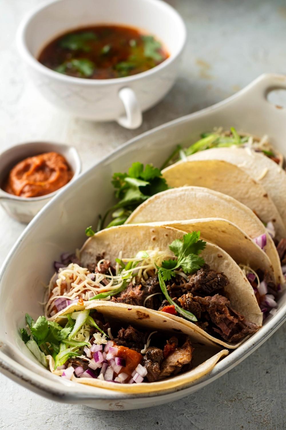 An oval serving bowl with a couple of Birria tacos in it. Behind it is a small bowl of dipping sauce and another bowl of consommé.