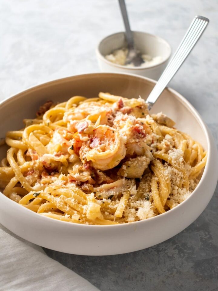 Part of a white bowl with chicken and shrimp carbonara in it. Or utensils submerged in pasta behind the ball is a small white bowl of Parmesan cheese.