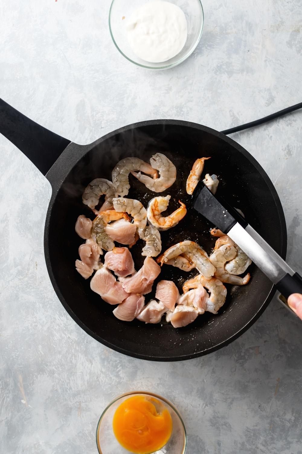 A skillet filled with chicken and shrimp. Behind it is above a girl going in front of it is a bowl of heavy cream.