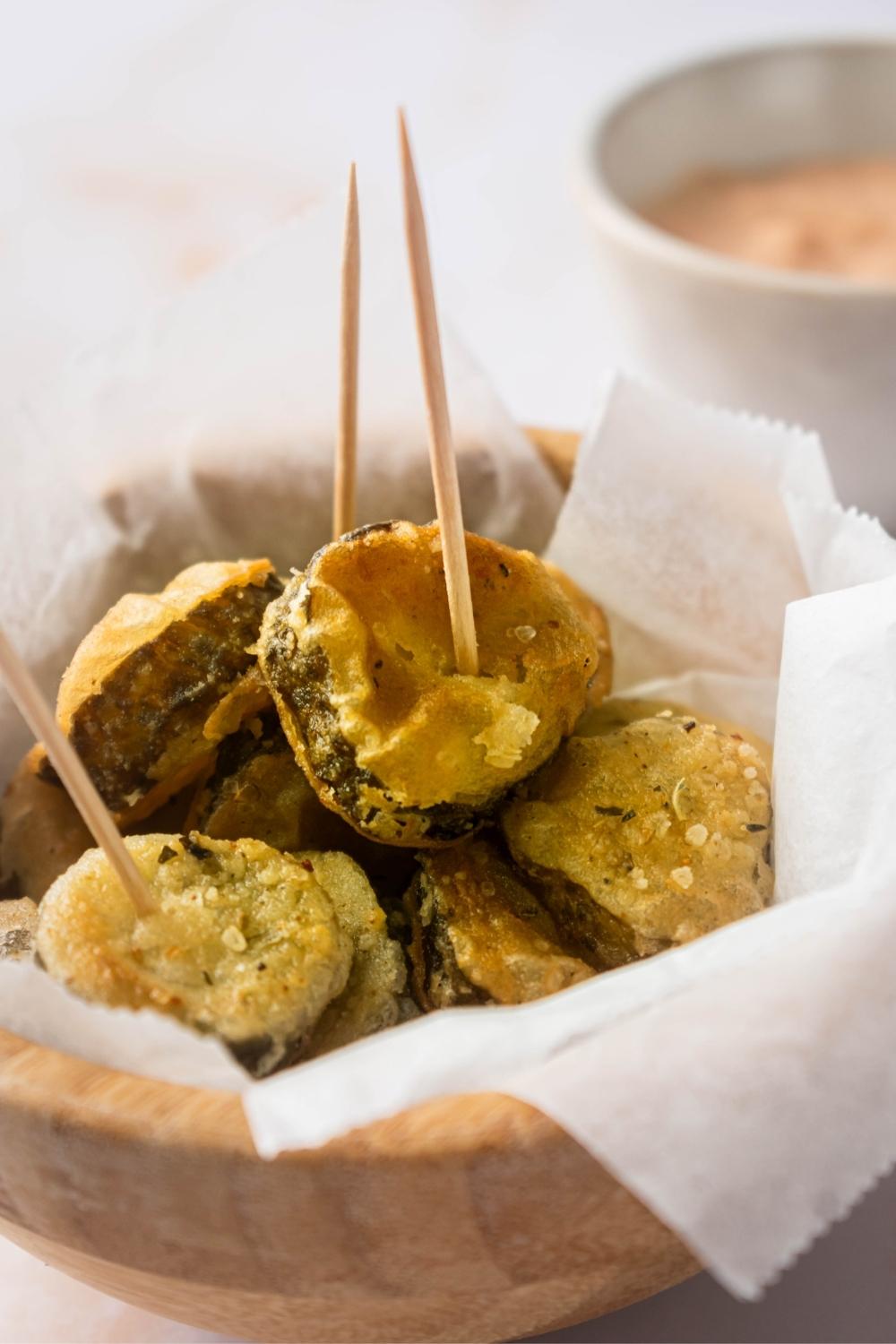 Fried pickle slices on parchment paper in a wooden bowl. The front to pickle slices have a toothpick in the center of them.