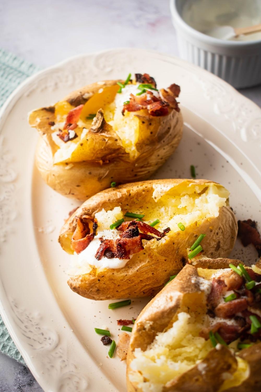 A white plate with three split open baked potatoes on it. The baked potatoes have some chives, sour cream, and bacon in them.