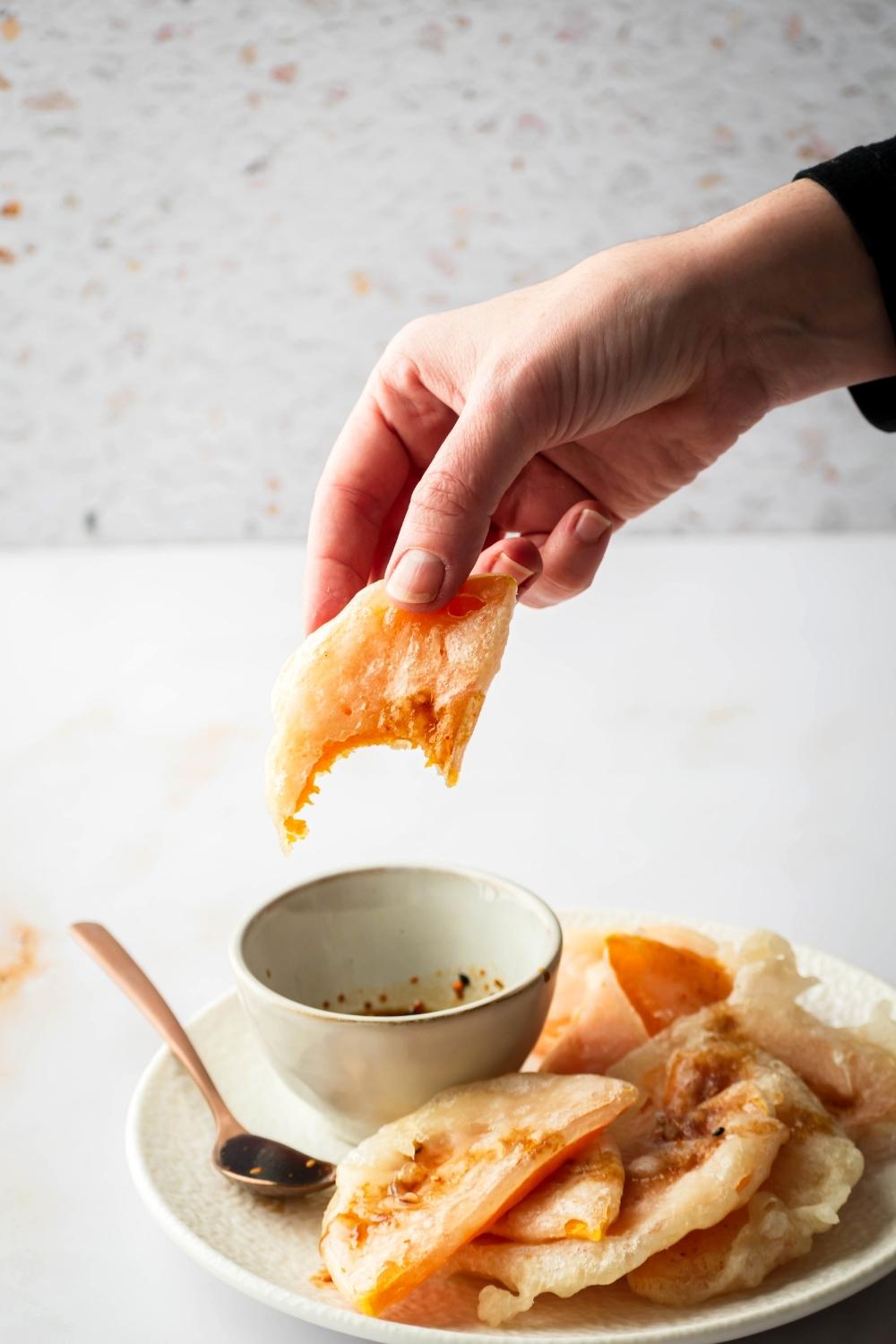 A hand holding a piece of fried squash that has a bite out of it over a cup of soy sauce that is on a white plate with a bunch of other pieces of fried squash overlapping with one another.