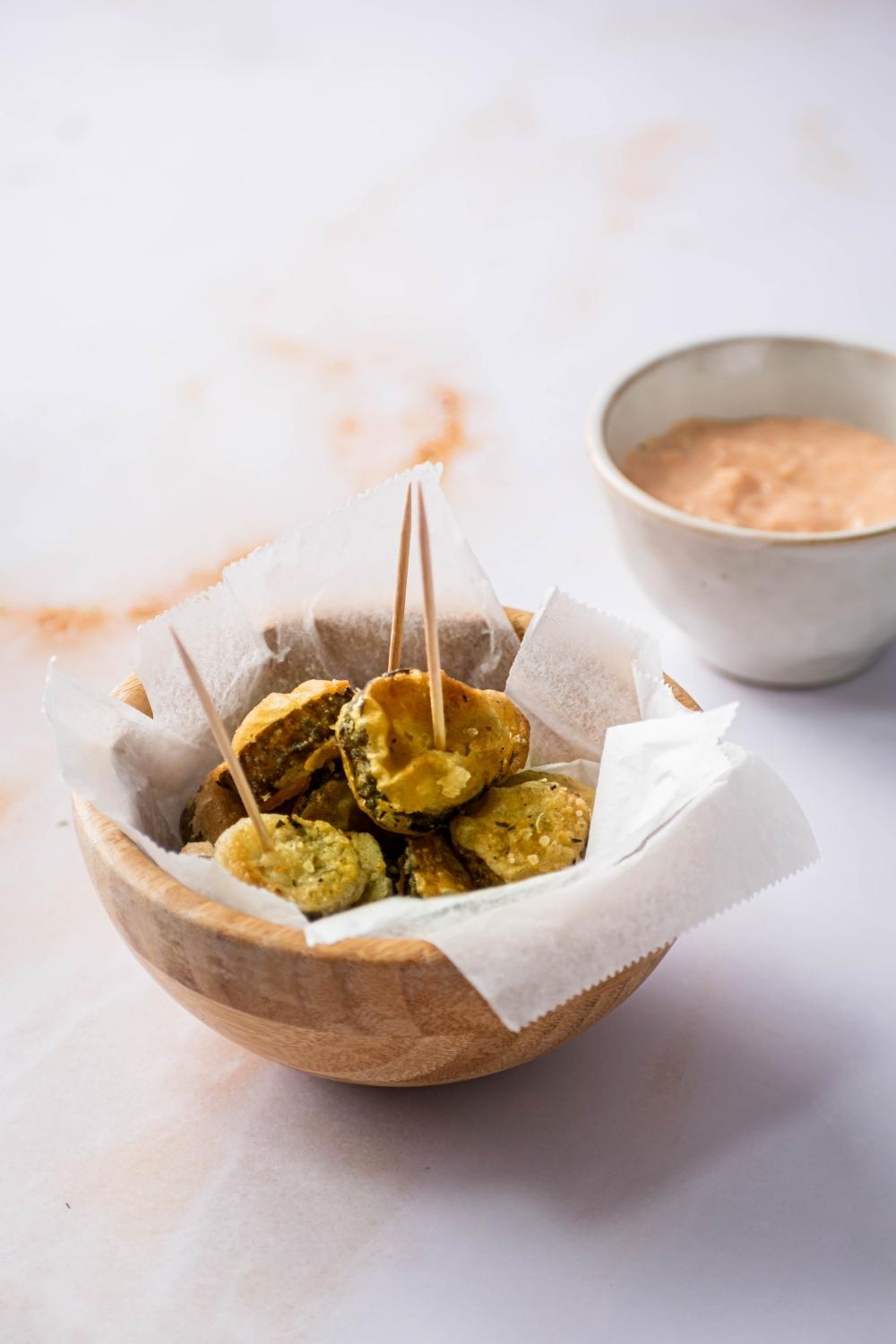 A bunch of fried pickle slices on parchment paper and a wooden bowl on a white counter. Behind the wooden bowl is a white bowl with a dipping sauce in it..