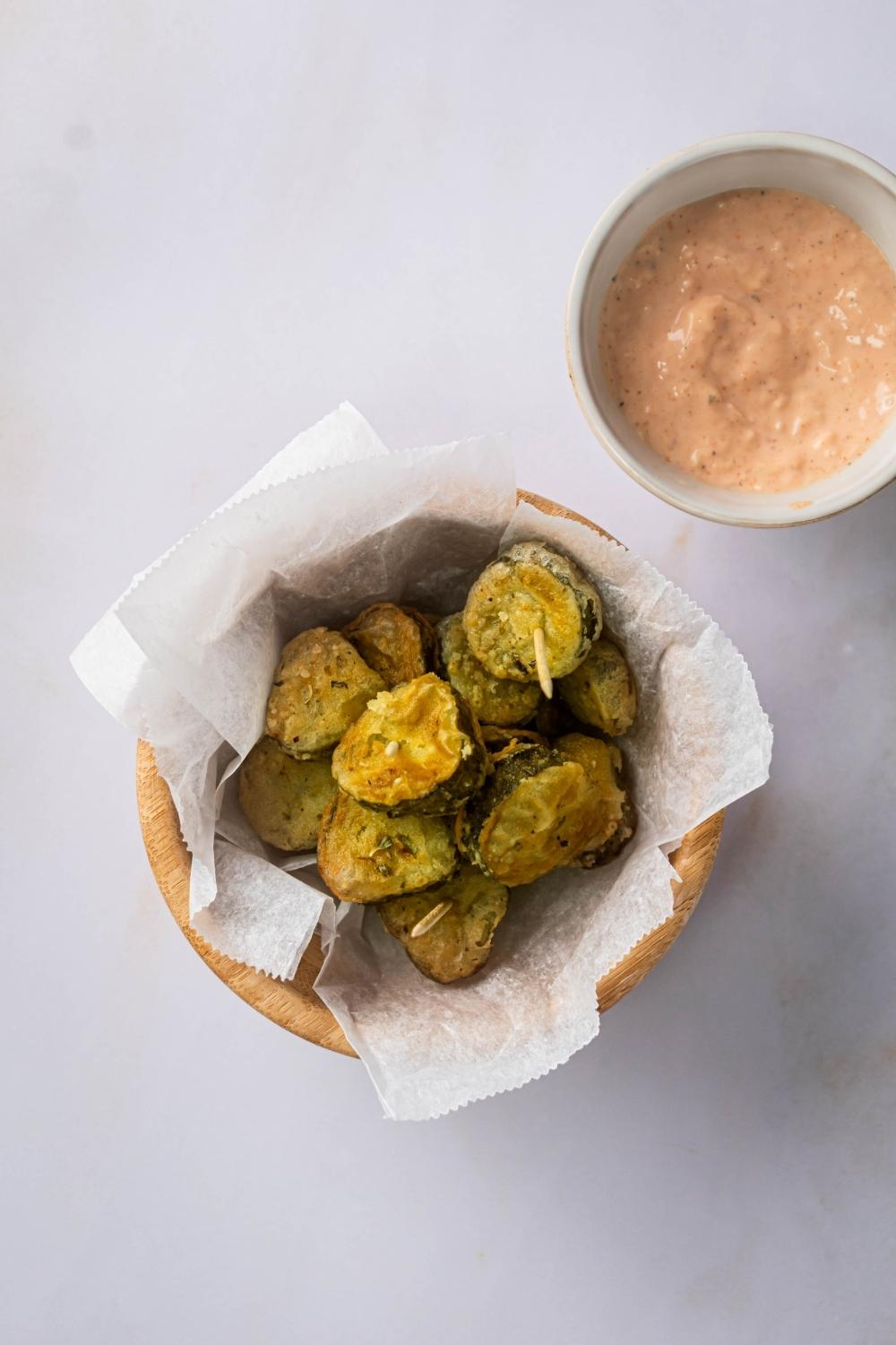 A bunch of fried pickle slices on top of a sheet of parchment paper that is in a wooden bowl on top of the white counter. Behind it is a white bowl with a dipping sauce in it.