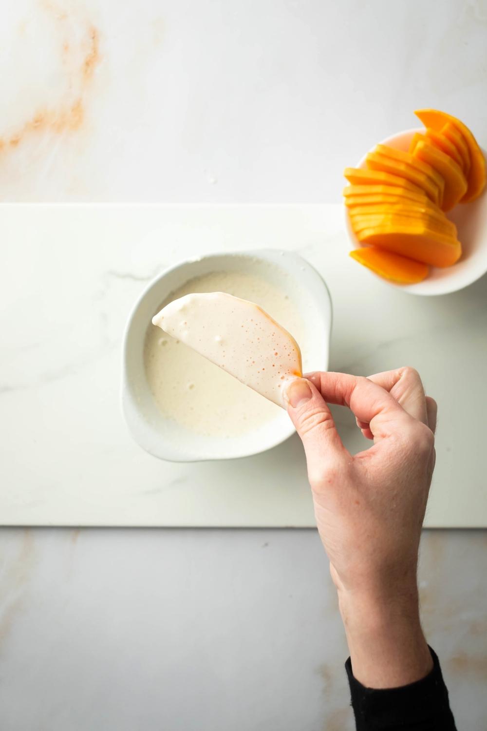 A hand holding a piece of squash coated in a flour mixture. It is being held over a bowl with the mixture in it and next to that bowl is a bowl of sliced squash.