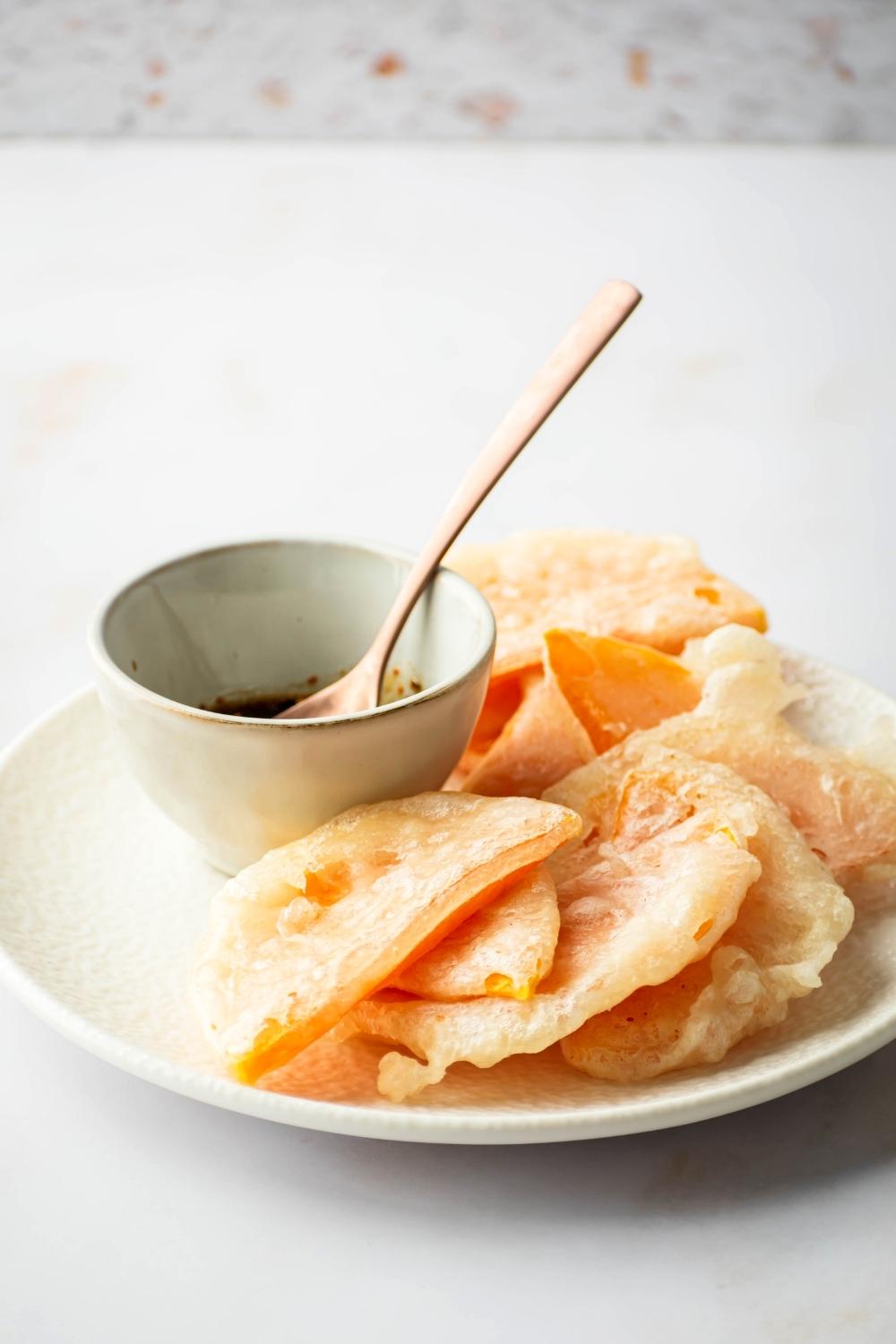 Slices of fried squash overlapping one another on a white plate with a bottle of soy sauce with a wooden spoon in it next to the squash on the plate.