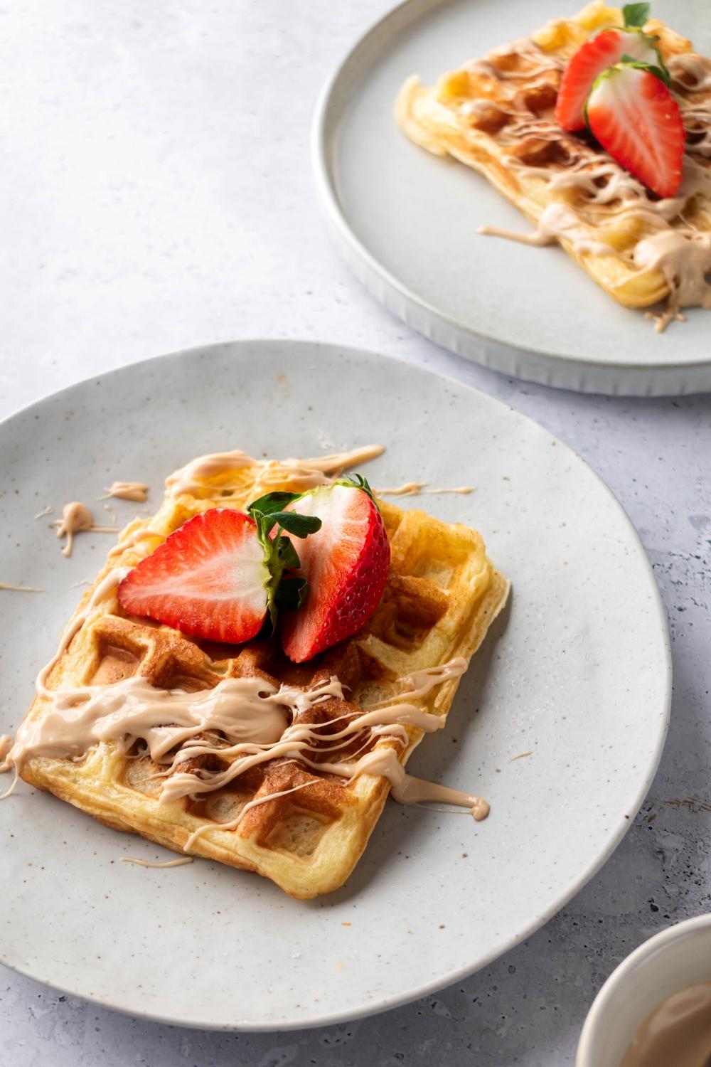 A waffle on top of a white plate with another waffle on a white plate behind it. Both waffles have a sliced strawberry on top and dolce de leche drizzled over it.