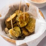 Several fried pickle slices on a sheet of parchment paper filled in a wooden bowl on the white counter. Behind that is part of the white bowl filled with a dipping sauce.