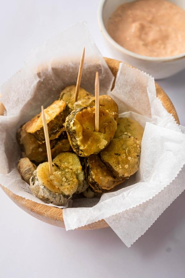 Several fried pickle slices on a sheet of parchment paper filled in a wooden bowl on the white counter. Behind that is part of the white bowl filled with a dipping sauce.