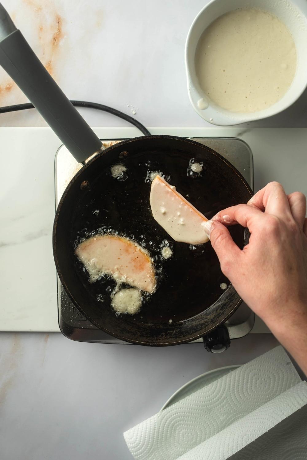 A hand holding a piece of fried squash over a frying pan filled with oil and one other piece of squash.