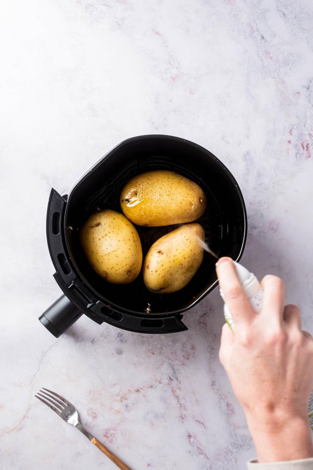Three potatoes in an air fryer. A hand is holding a bottle of cooking spray and spraying it on the potatoes.