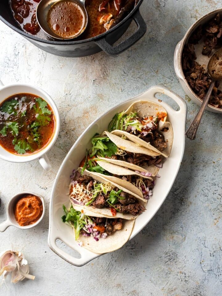 A white serving bowl filled with six Birria tacos. Next to it to the left is a small bowl of dipping sauce, behind that a white bowl of consomme, behind that part of a dutch oven with the consomme ingredients in it.