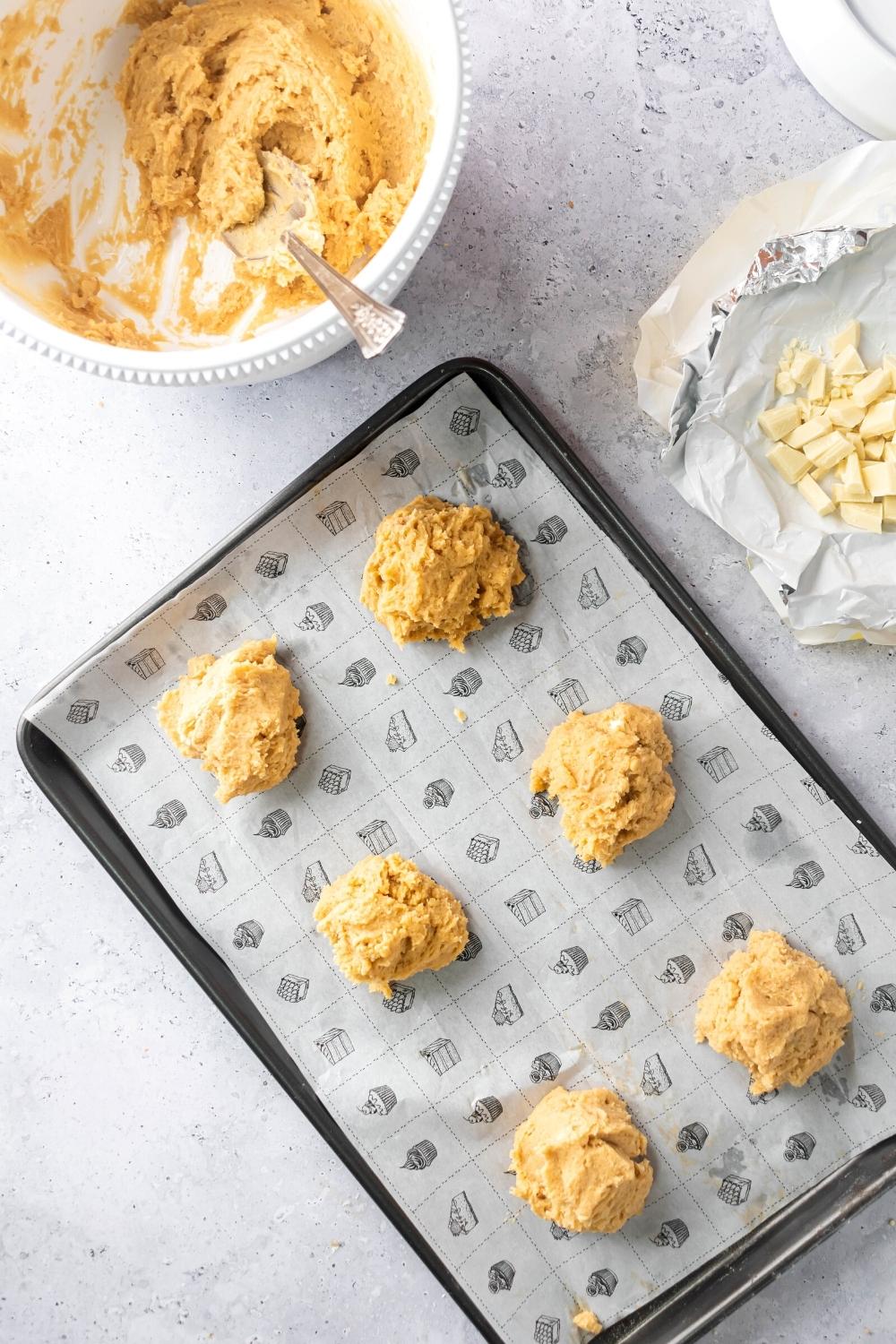 Six cookie dough balls on a baking sheet lined with parchment paper. Behind it as part of a bowl with white chocolate chunks in it and part of another bowl with the rest of the cookie dough in it.