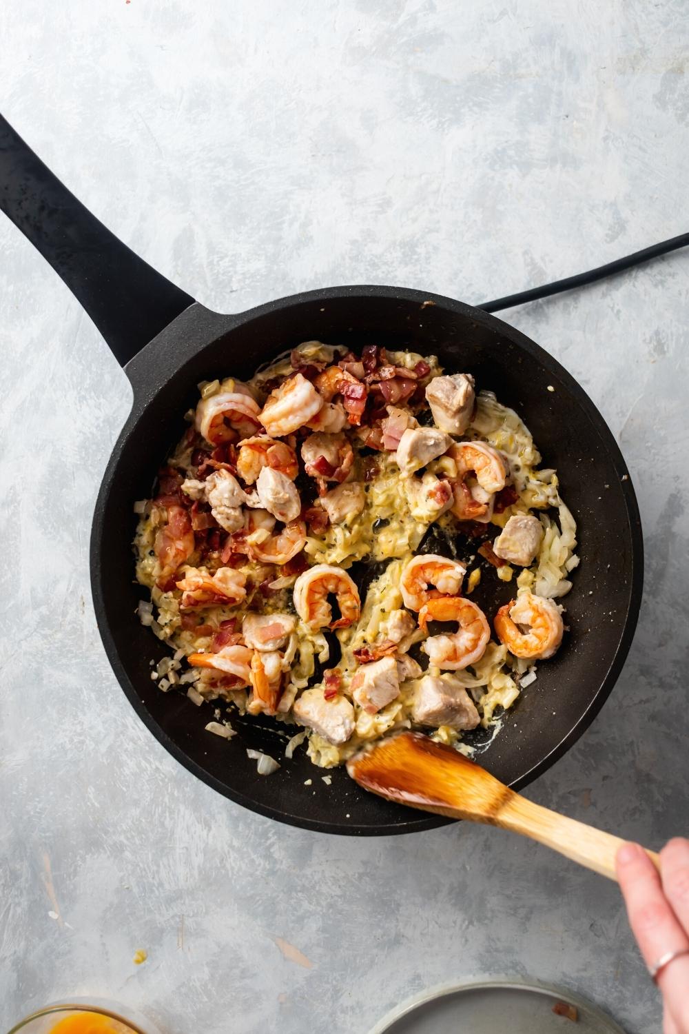Hey skillet filled with chicken, shrimp, bacon, onion, and garlic. A wooden spoon is stirring everything around.