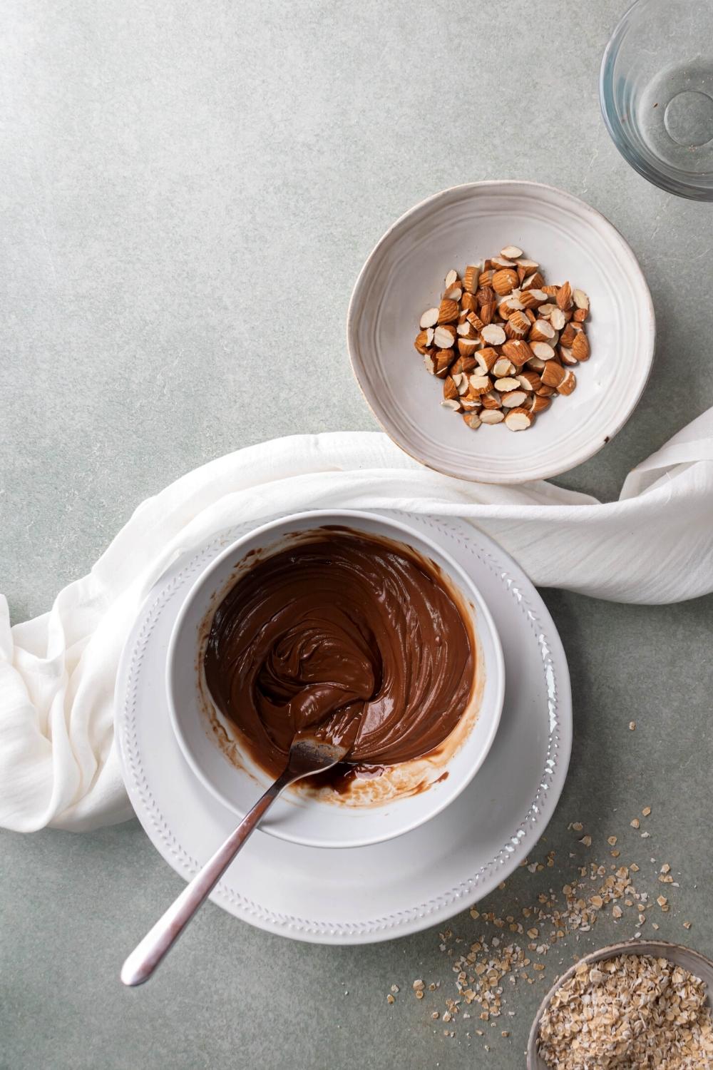 A white bowl filled with chocolate sauce with the spoon in it with a bowl of chopped almonds behind it on the gray counter.
