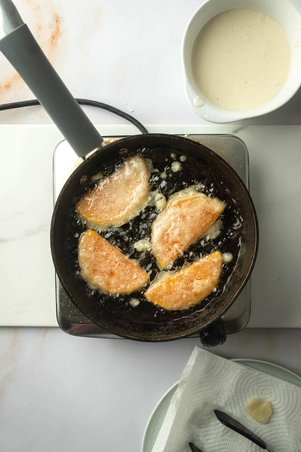 A frame pan filled with oil and four pieces of squash. The frying pan is on a square burner and behind is a bowl thee mixture for breading.
