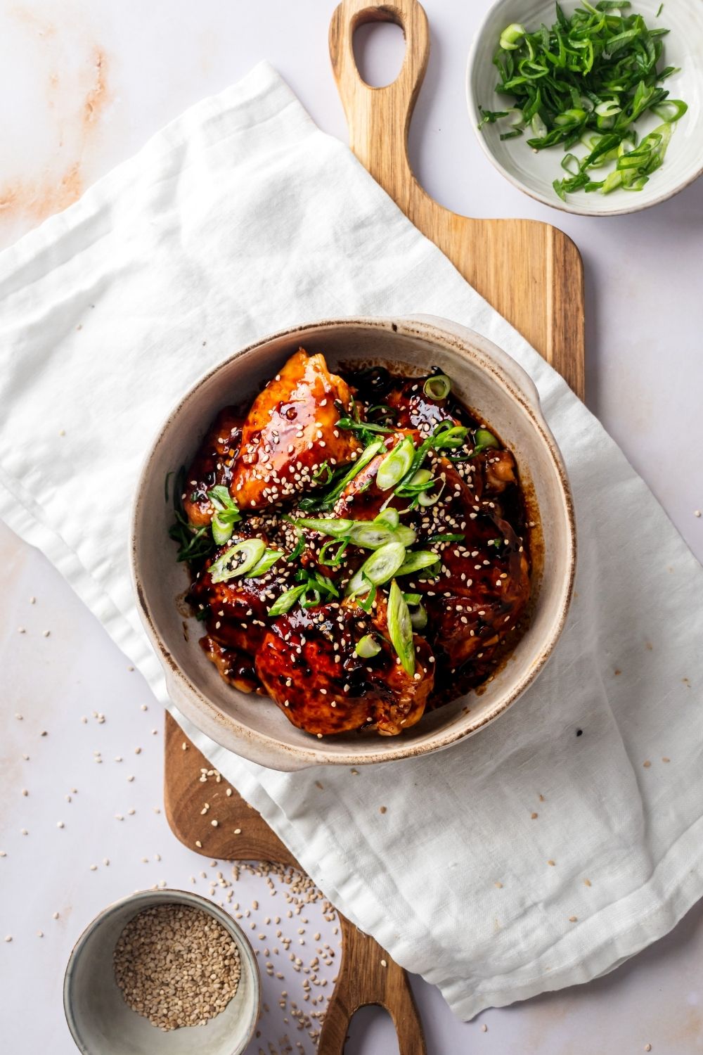 A couple of huli huli chicken thighs in a white bowl that is on top of a white table cloth on a wood cutting board on a white counter.
