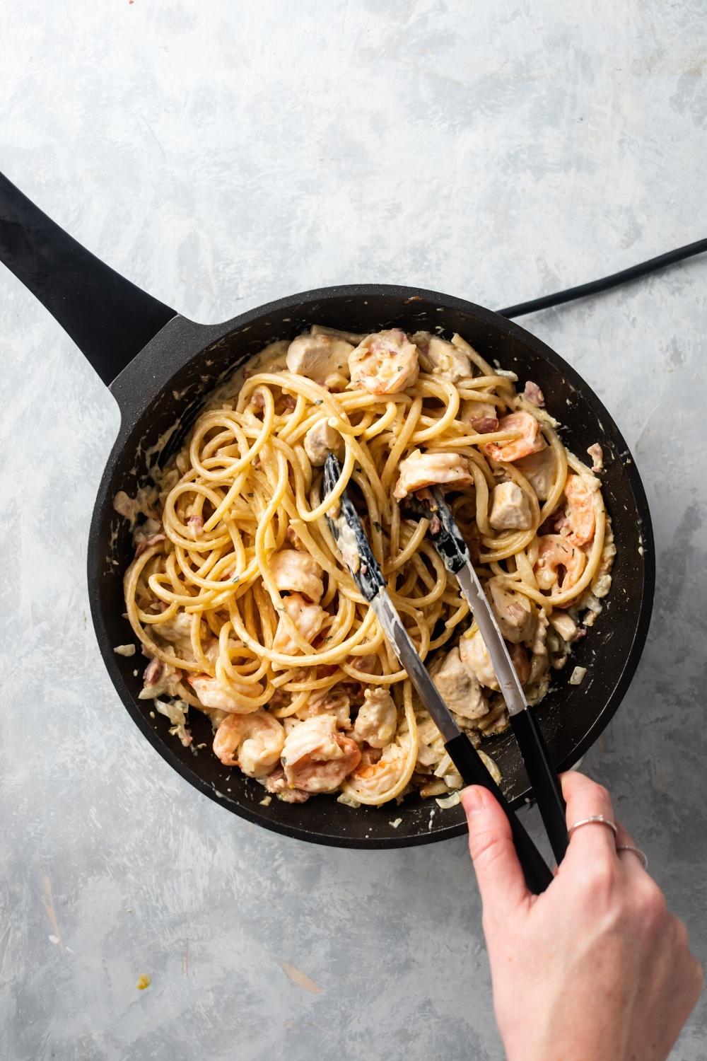 Hey skillet filled with chicken and shrimp carbonara. A hand is holding tongs in the pasta.