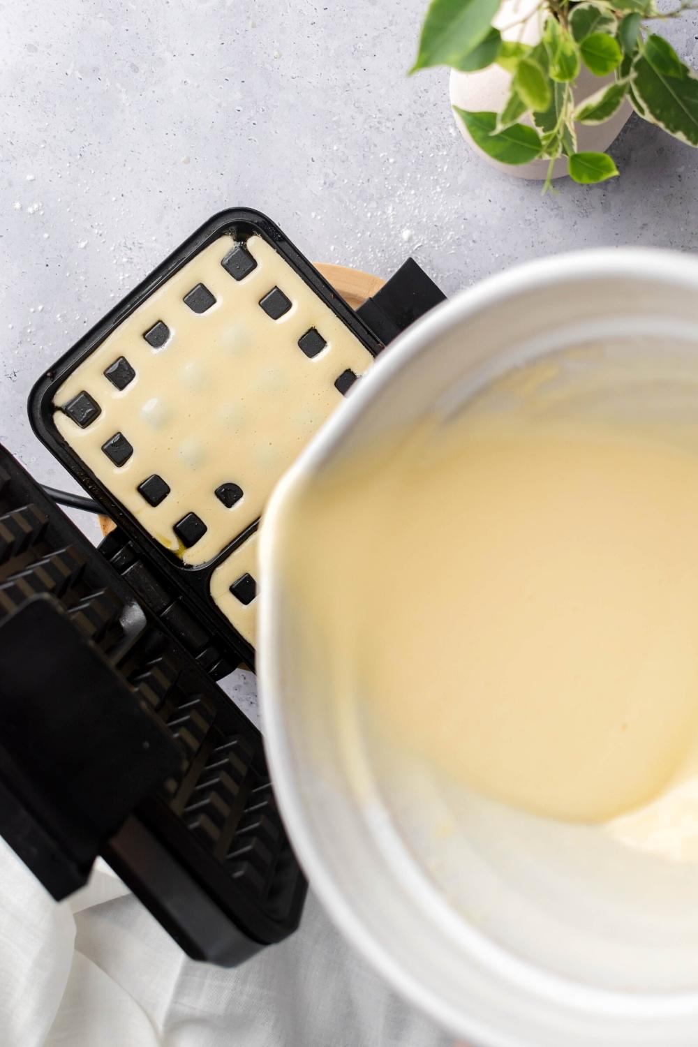 A white bowl with some waffle batter in it pouring some of the batter into an opened waffle iron. The waffle iron is filled with the batter.