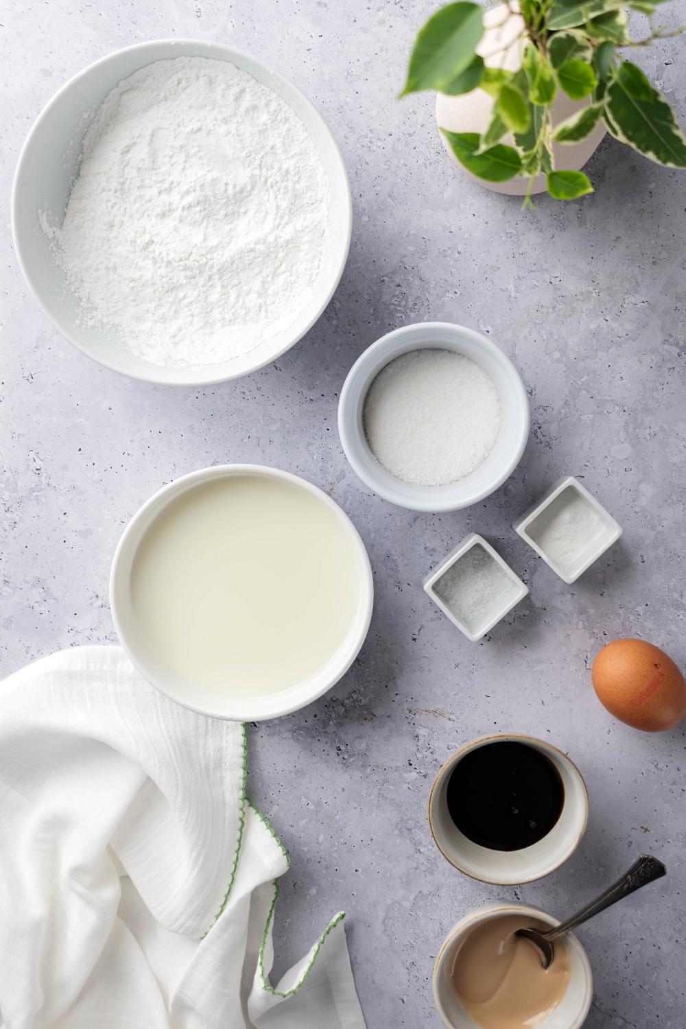 A bowl of sweet rice flour, a bowl of sugar, a bowl of milk, an egg, and a bowl of baking powder all on a gray counter.