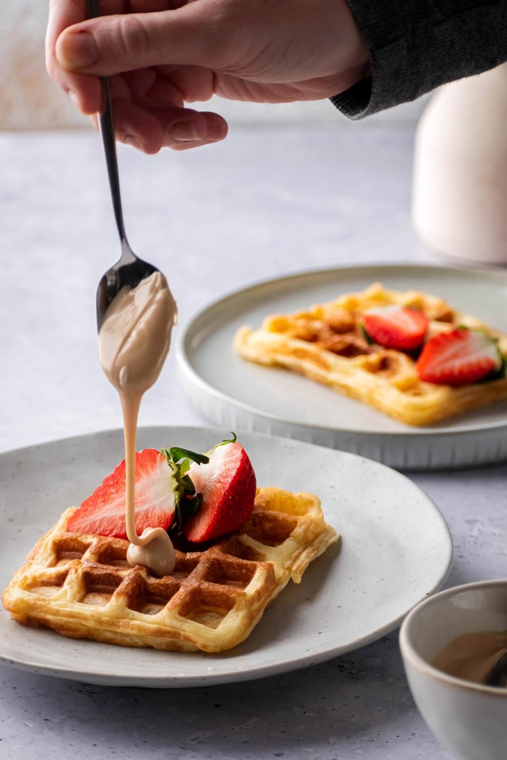 A hand holding a spoon with dulce de leche on it drizzling it over the top of a waffle that is on a white plate. Behind it is part of another white plate with a waffle on it.
