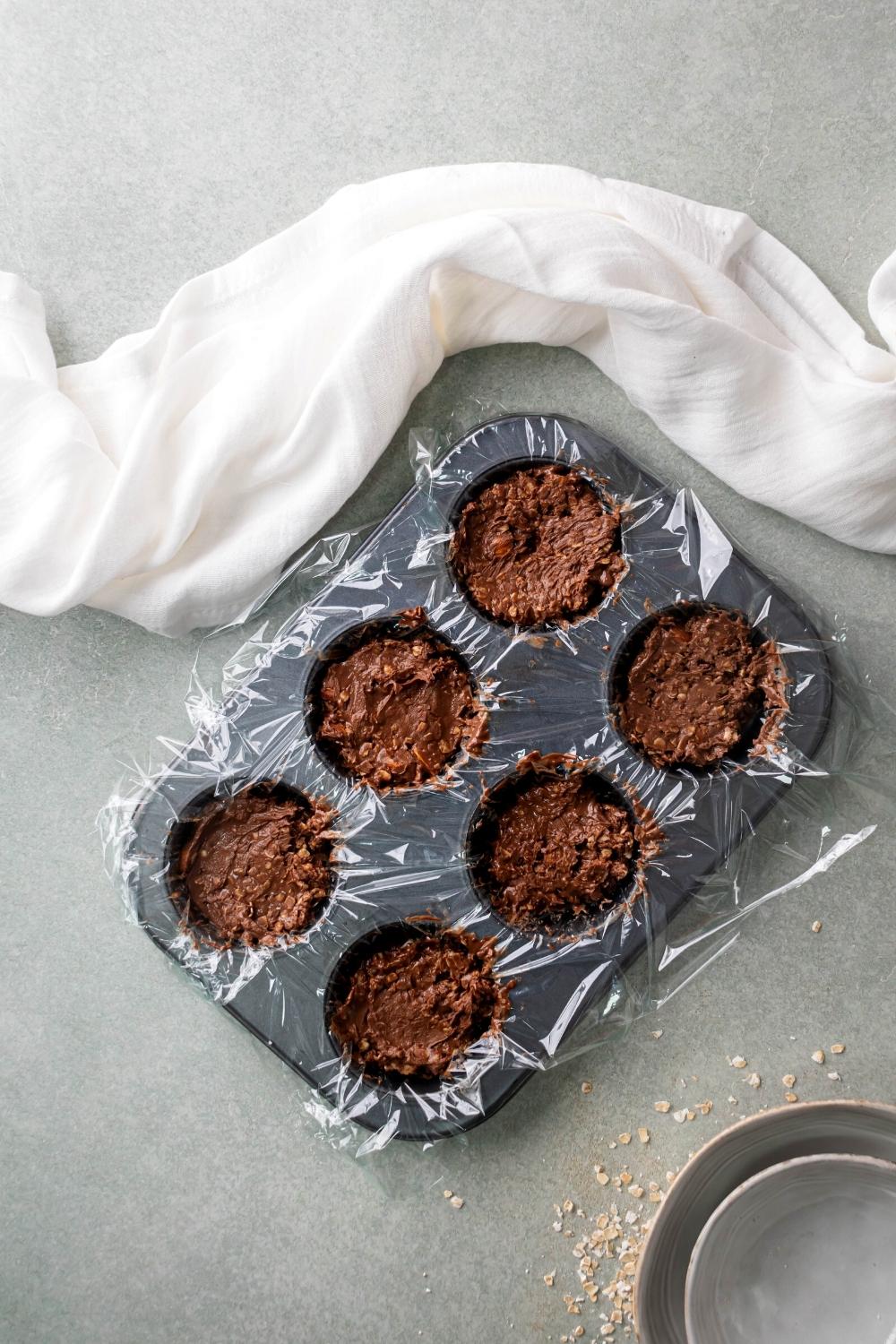 A six slotted mini muffin pan line with plastic wrap filled with no bake chocolate oatmeal cookies.