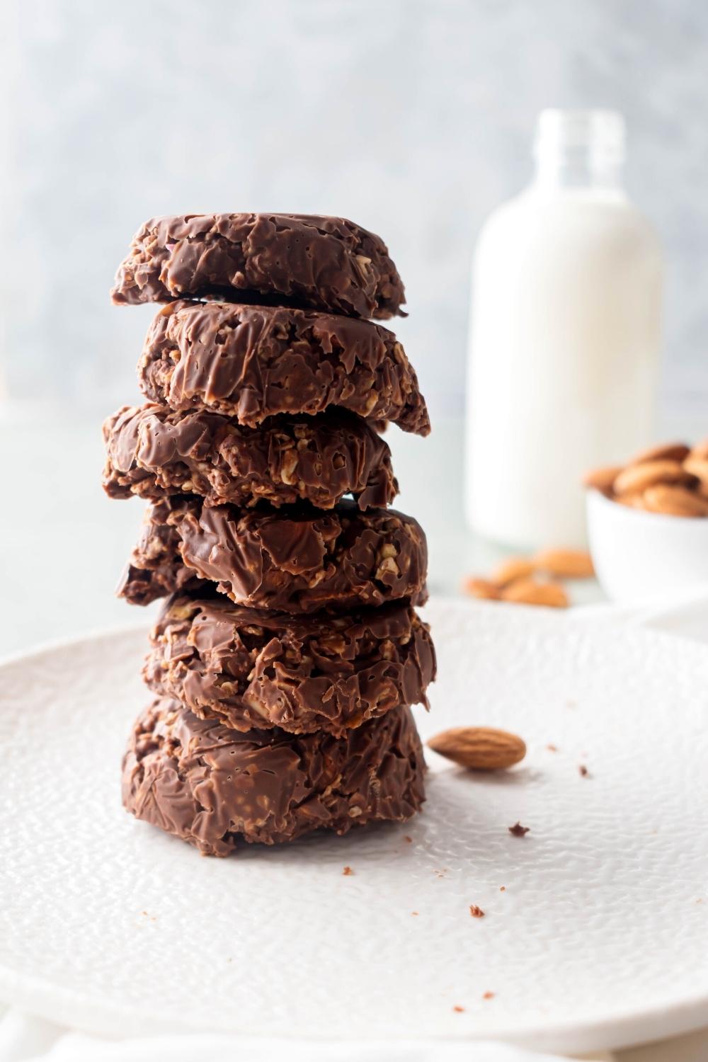 Six no bake chocolate oatmeal cookies stacked on top of one another on a white plate. There is a glass jar filled with milk behind them.