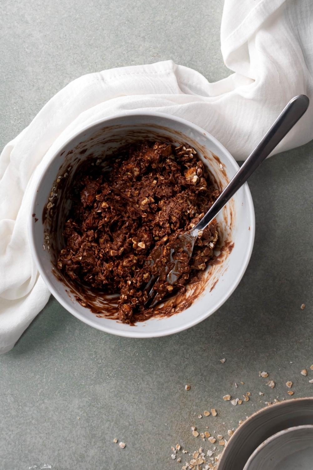 A white bowl filled with chocolate oatmeal no bake cookie. A fork is submerged in the mixture and the bowl is on a grey counter.
