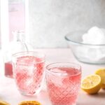 Two glasses filled with pink lemonade vodka and a few ice cubes on a white counter. Also on the counter around the glasses are lemon slices, a bowl of ice, a glass jar of pink lemonade, and a bottle of vodka.